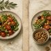 Vista superior de dos platos de ensalada mediterránea con garbanzos, tomates y hojas verdes; en uno se vierte aceite de oliva y en el otro aparece un cuenco con frutos secos.