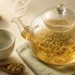Tetera de vidrio con infusión de manzanilla y flores visibles, acompañada de una taza, flores secas y una cuchara de madera.