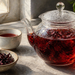 Tetera de vidrio con infusión roja de hibisco junto a una taza y un cuenco con flores secas sobre una superficie clara cerca de una ventana.