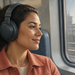 Mujer adulta de cabello oscuro y rizado escucha música con auriculares grandes mientras viaja sentada junto a la ventana de un tren y observa el paisaje urbano.