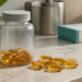 Frasco transparente con cápsulas doradas de omega-3 y varias cápsulas sueltas sobre una encimera clara.