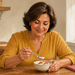 Mujer adulta sonriente comiendo un cuenco de yogur con fresas, sentada a una mesa de cocina mientras sostiene una cuchara con una pequeña porción.