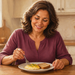 Mujer adulta sonriendo mientras come huevos revueltos y una tostada de pan integral, sentada a una mesa en una cocina luminosa.