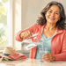 Mujer adulta sirviendo agua de una jarra de vidrio en un vaso dentro de una cocina luminosa, con frutas frescas y plantas alrededor.
