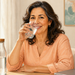 Mujer madura sonriente bebe agua de un vaso transparente, sentada a una mesa con una botella de agua en una habitación luminosa.