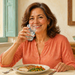 Mujer madura sonriente sostiene un vaso de agua junto a la boca mientras come pescado con verduras en una mesa junto a una ventana luminosa.