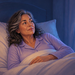 Mujer madura despierta en la cama durante la madrugada, mirando hacia la ventana mientras permanece recostada entre almohadas y sábanas blancas.