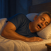 Hombre mayor duerme de lado con la boca abierta sobre una almohada, en un dormitorio nocturno iluminado por una lámpara junto a la cama.