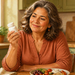 Mujer mayor observando un frasco blanco de suplementos mientras está sentada ante un plato con verduras, legumbres y otros alimentos en una cocina luminosa.