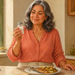 Mujer mayor sonriente sostiene un pequeño salero de cristal junto a un plato de pescado a la plancha con verduras, en una cocina luminosa.