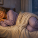 Mujer mayor con pijama lila duerme de lado, con la cabeza apoyada sobre una almohada y las piernas flexionadas bajo un edredón blanco.
