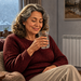 Mujer mayor con jersey burdeos sostiene un vaso de agua mientras descansa en un sofá, junto a un radiador encendido en una sala de estar durante el invierno.