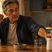 Hombre mayor fumando junto a un cenicero, un vaso de agua y un frasco de suplementos sobre la mesa.