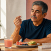 Hombre mayor sosteniendo un comprimido junto a un vaso de zumo de pomelo, medio pomelo y un pastillero sobre la mesa.