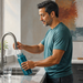 Hombre adulto llena una botella reutilizable con agua del grifo en la cocina, junto a un vaso de agua sobre la encimera.