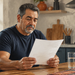 Hombre adulto de cabello canoso sentado en una cocina leyendo un documento, con un vaso de agua sobre la mesa.