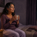 Mujer mayor sentada en la cama por la noche sosteniendo una taza, con un frasco y hierbas secas sobre la mesita de noche.