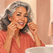 Mujer mayor de cabello gris y rizado sonríe mientras utiliza hilo dental entre los dientes frente al lavabo de su cuarto de baño.