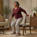 Mujer mayor realizando una sentadilla asistida junto a una silla en el salón de casa, con una mano apoyada en el respaldo para mantener el equilibrio.