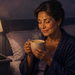 Mujer adulta sentada en la cama por la noche mientras sostiene una taza humeante con ambas manos junto a una lámpara encendida.