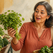Mujer adulta en un mercado sosteniendo y observando un manojo de berros, rodeada de espinacas, brócoli y otras verduras de hoja verde.