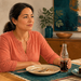Mujer sentada a la mesa con una botella de refresco y un trozo de pastel junto a un plato vacío en un comedor luminoso.