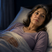 Mujer adulta de cabello castaño con canas permanece despierta en la cama durante la noche, con expresión pensativa y la mirada dirigida hacia un lado, mientras la luna ilumina la ventana.