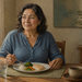Mujer adulta sentada a la mesa comiendo pescado con arroz, brócoli y verduras, acompañada de un vaso de agua y una taza.