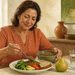 Mujer adulta sentada a la mesa sirve lentejas en su plato, acompañadas de verduras, pan, una pera y un vaso de agua, en una escena relacionada con una alimentación rica en fibra.