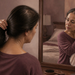 Mujer adulta frente al espejo del dormitorio recogiendo su cabello con una mano mientras observa su pelo y cuero cabelludo sobre un tocador con cepillo y productos de cuidado personal.