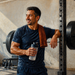 Hombre en un gimnasio junto a una barra con discos, sosteniendo una botella de agua y descansando con una toalla sobre el hombro después del entrenamiento de fuerza.