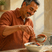 Hombre mayor en una cocina añade semillas de linaza a un bol con avena, yogur y frutos rojos, junto a otros alimentos sobre la encimera.