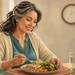 Mujer mayor sentada a la mesa comiendo un plato con proteína, cereales y verduras junto a un vaso de agua, con un reloj visible al fondo.