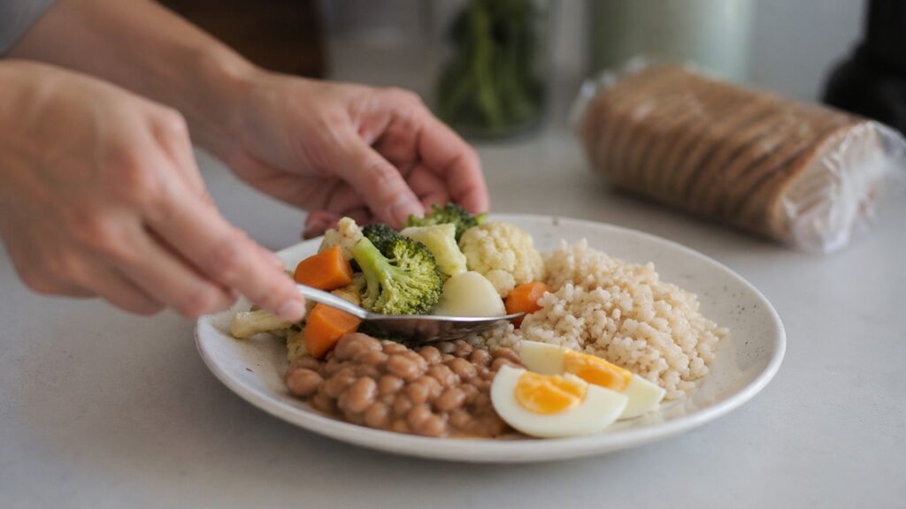 Mãos montando uma refeição simples com feijão, legumes, ovo e arroz integral.